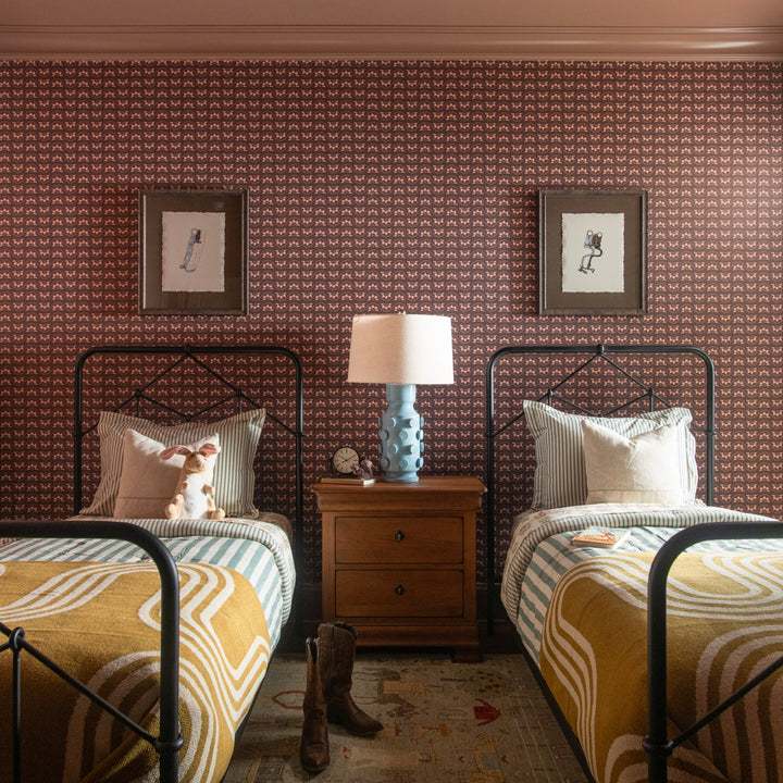 Two beds with patterned bedding and gold throw blankets in a room with decorative wallpaper and a lamp.