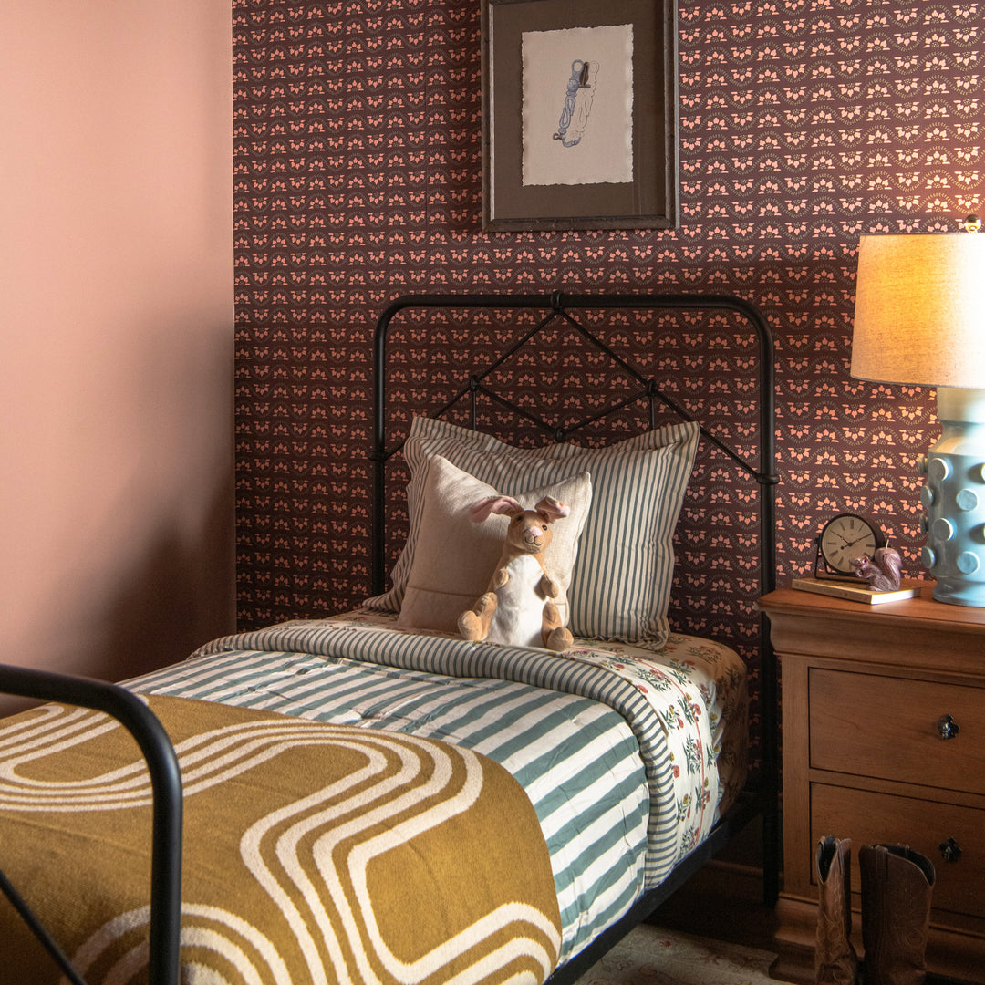 Bedroom with patterned wallpaper, bed with striped bedding, and modern gold throw blanekt and nightstand with lamp.