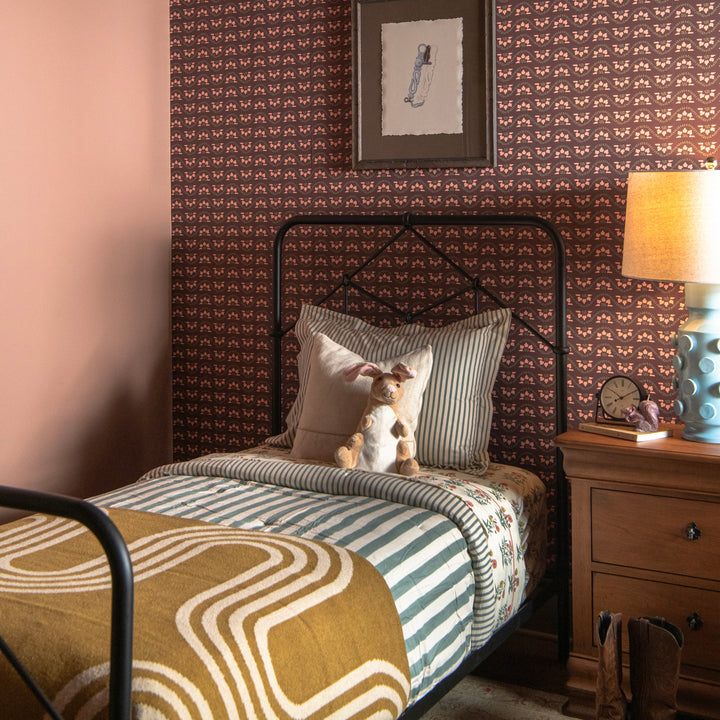 Bedroom with patterned wallpaper, bed with striped bedding, and modern gold throw blanekt and nightstand with lamp.