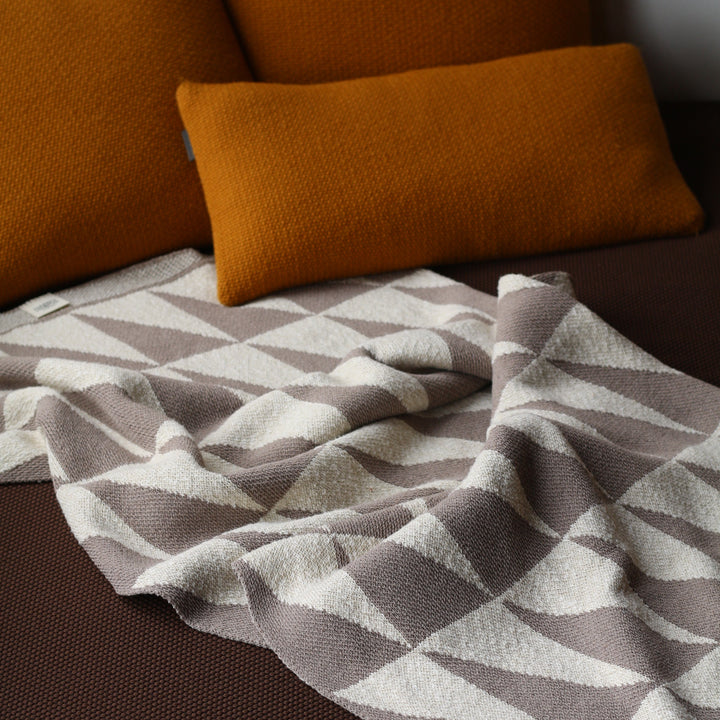 Taupe and ivory patterned blanket draped over a brown surface with orange pillows.