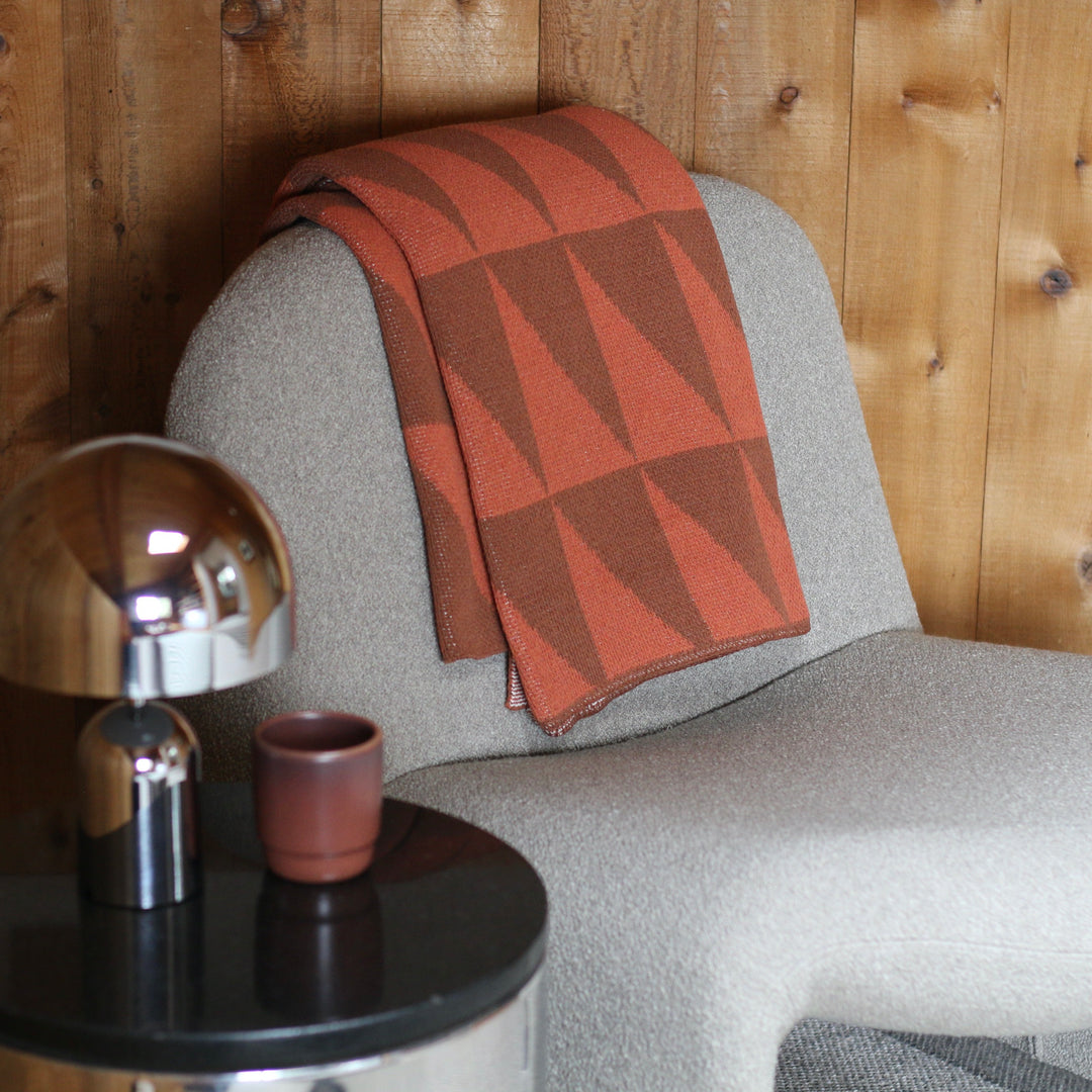 cream armchair with a orange patterned blanket, small table with lamp and cup against wooden wall.