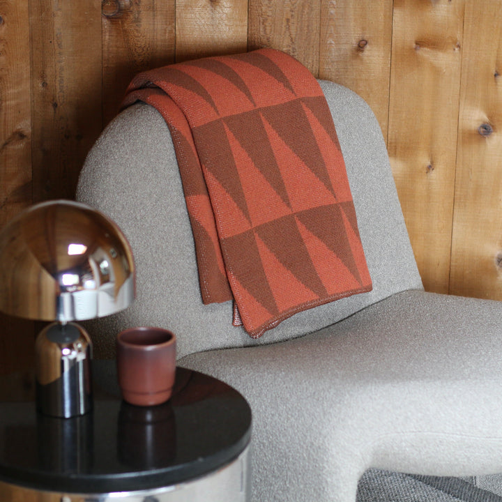 cream armchair with a orange patterned blanket, small table with lamp and cup against wooden wall.
