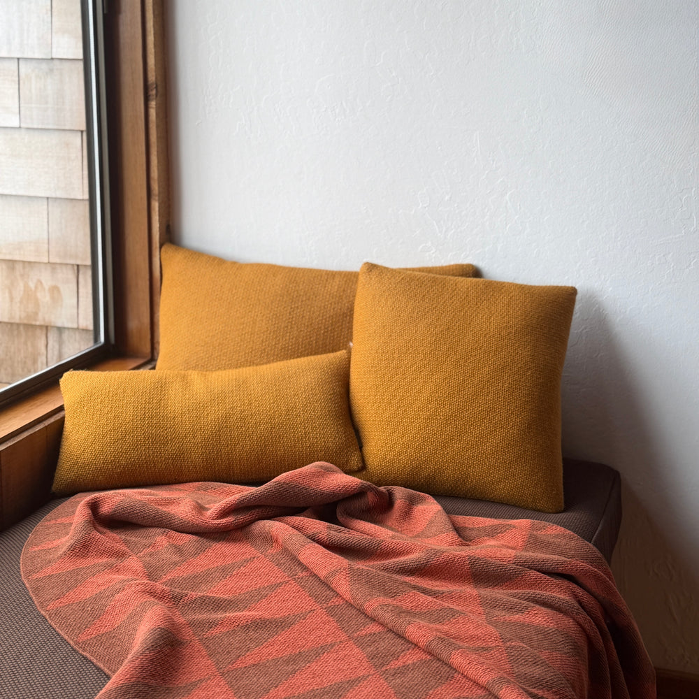 Mustard yellow pillows on a sofa with a orange patterned blanket, near a window.
