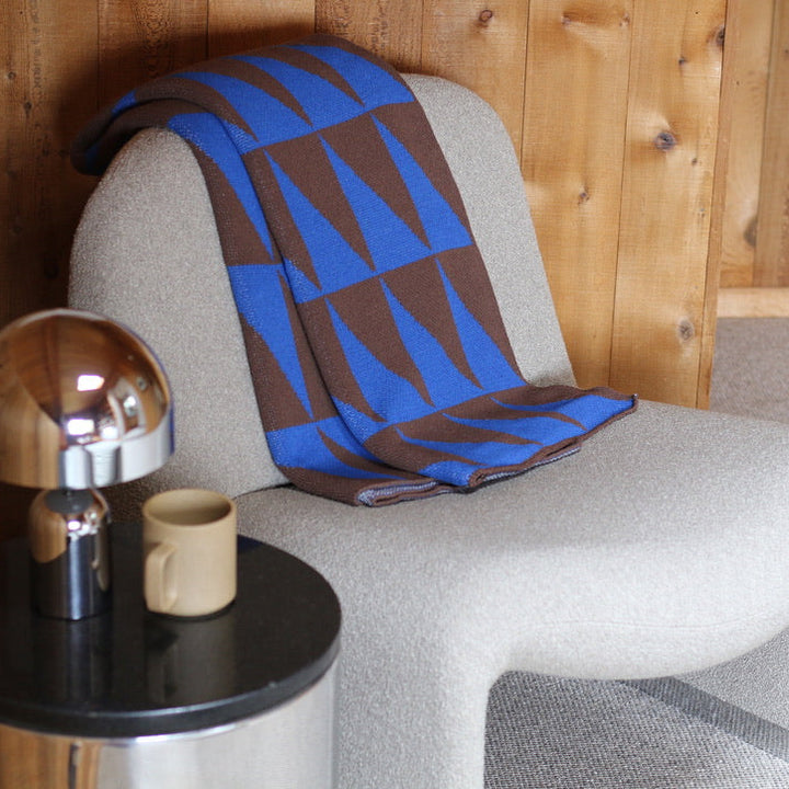 Cream chair with a blue and brown patterned blanket, next to a small round table with a lamp and mug.