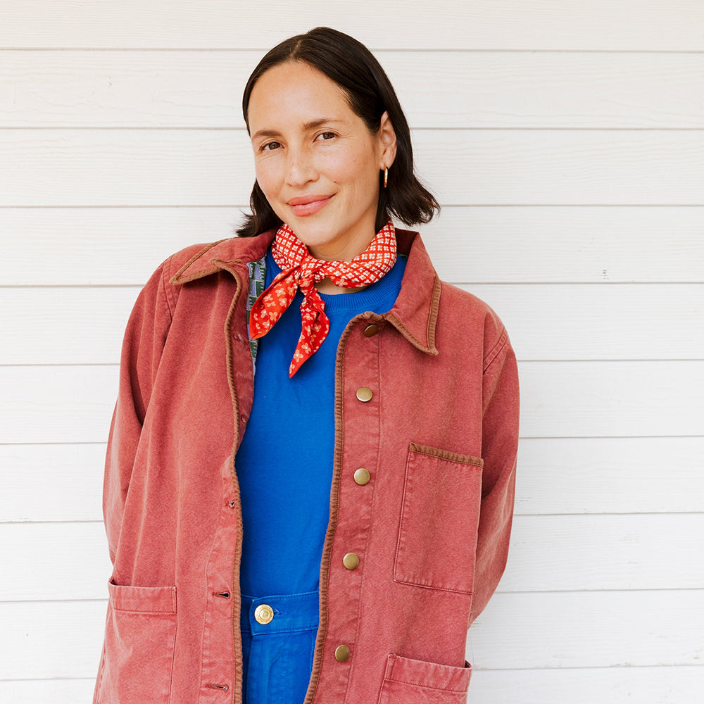Woman wearing a red jacket and blue shirt with a patterned scarf, standing against a white wooden background.