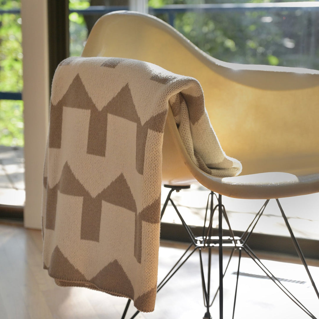 A cream and taupe knit throw blanket with a geometric house pattern draped over a modern molded chair in a bright room with a large window and green foliage outside.
