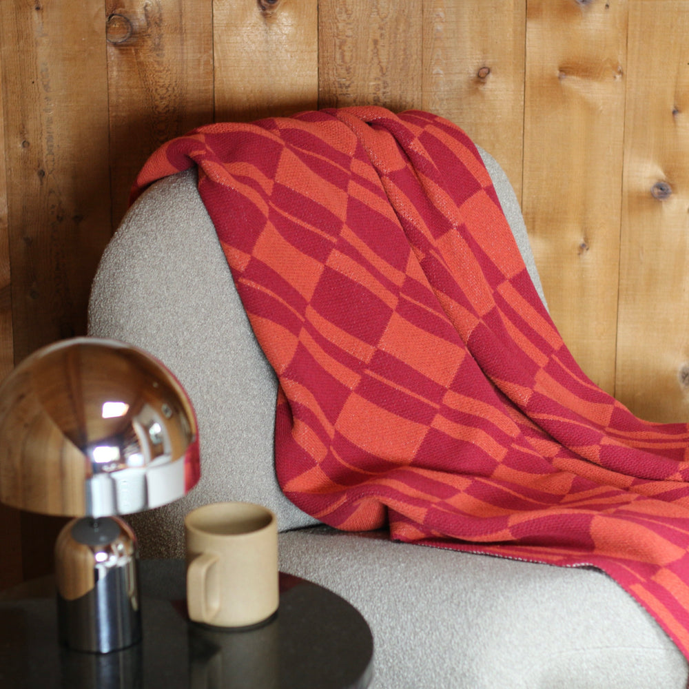 Red checkered blanket draped over a cream chair with a metallic lamp and beige mug on a wooden surface.