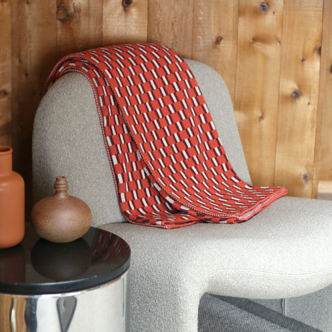 Red and orange patterned blanket draped over a gray chair with wooden wall and small table in the foreground