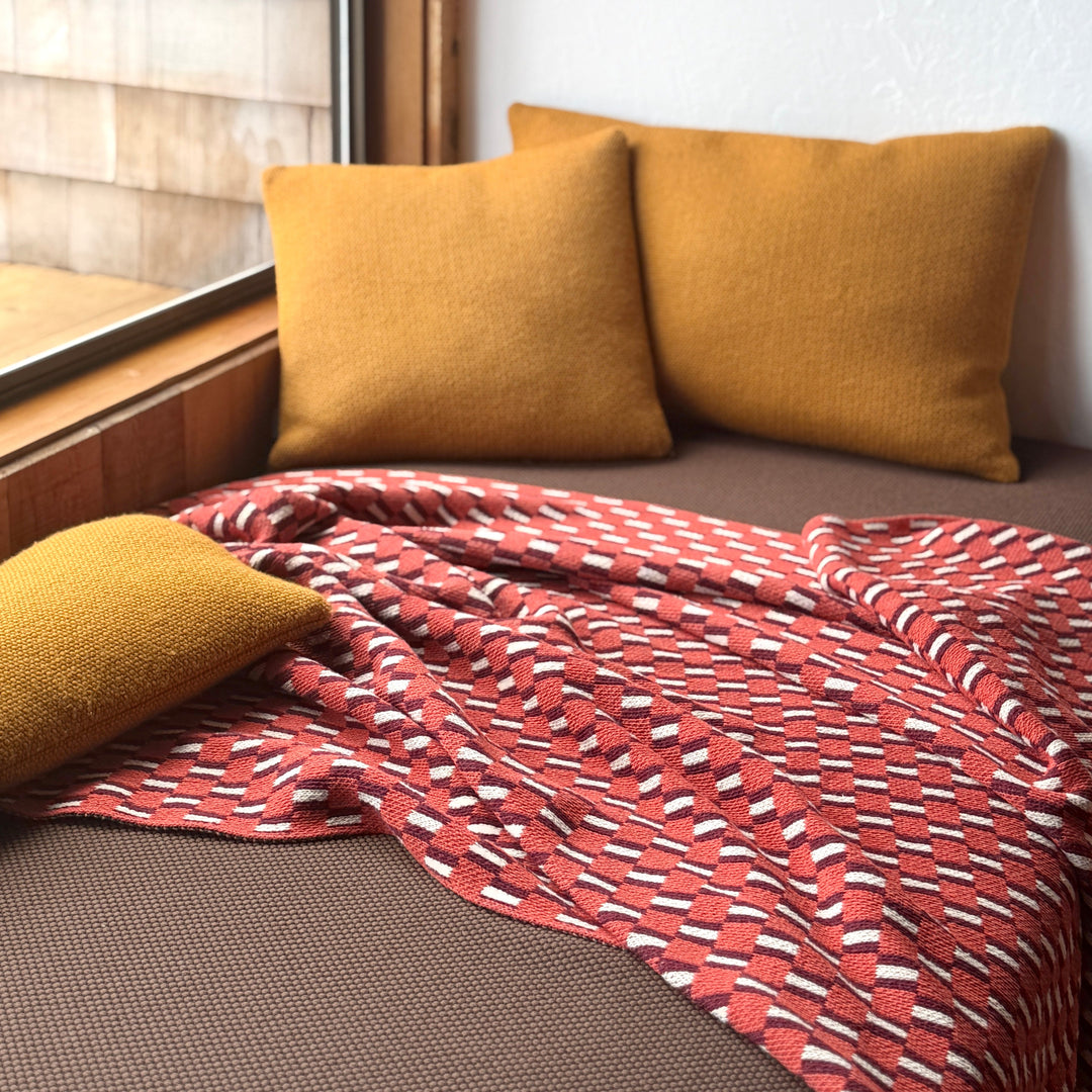 Two mustard yellow pillows on a bed with a red patterned blanket near a window.
