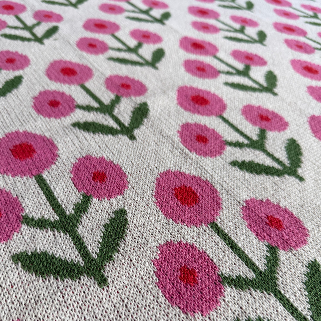 Close-up of a knitted textile with a repeating floral pattern of pink flowers and green stems on a white background.