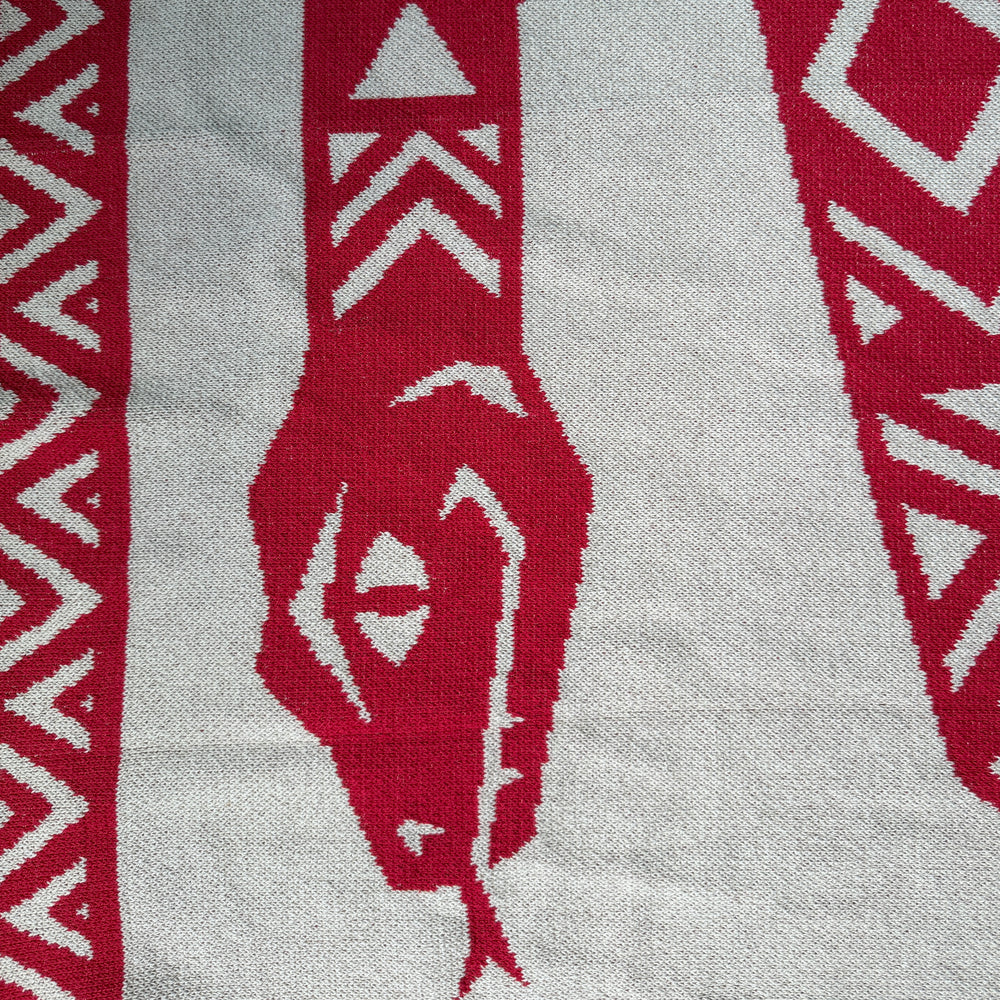A close-up of a knitted textile with a bold red and beige geometric pattern, featuring stylized tribal or animal-inspired motifs.
