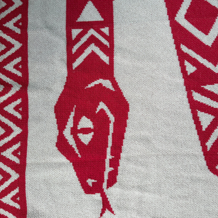A close-up of a knitted textile with a bold red and beige geometric pattern, featuring stylized tribal or animal-inspired motifs.