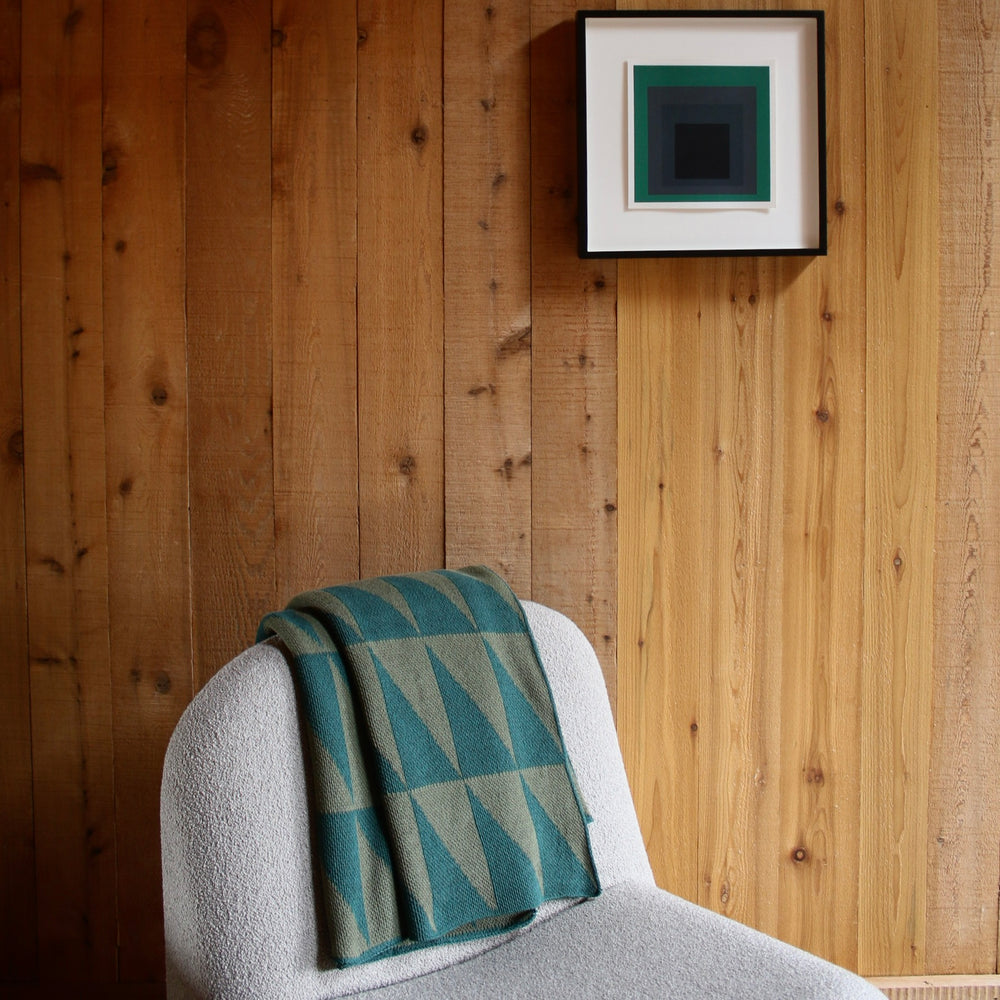 cream chair with a green triangle patterned blanket against a wooden wall with a framed picture