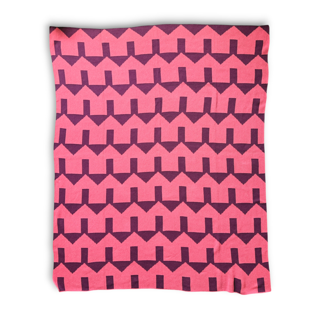 a throw blanket featuring a bold geometric house pattern of coral and dark red zig-zag stripes, shown flat against a white background.