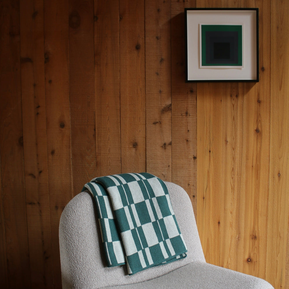 Green checkered blanket on a cream chair against a wooden wall with a framed picture.