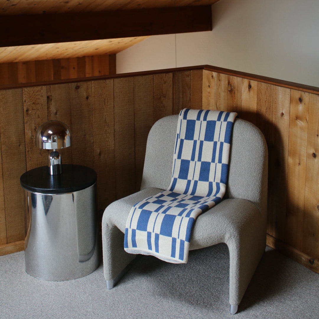 cream chair with a blue and white checkered blanket next to a metallic side table in a room with wooden paneling.