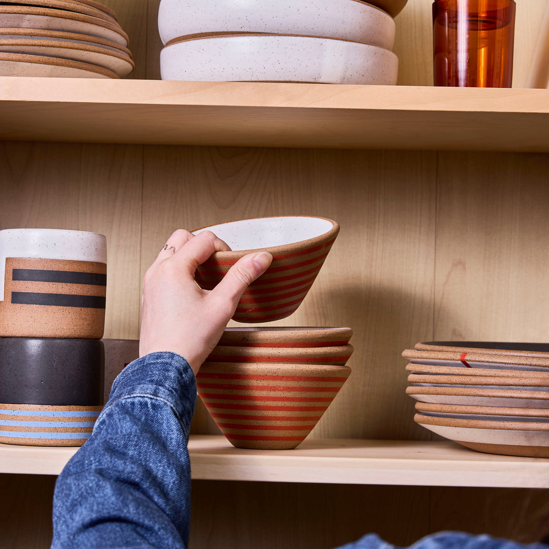 Person selecting a ceramic bowl from a wooden shelf with various bowls and cups.